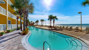 Ein Pool mit geschwungener Form, umgeben von Palmen und Liegestühlen, mit Blick auf das Meer und einem gelben Gebäude im Hintergrund.
