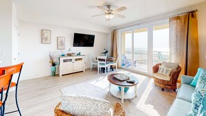 A spacious living room with a ceiling fan, a television, a dining area, and a balcony with a view. - Ocean-view 10th-floor condo with balcony, heated outdoor pool, hot tub, gym, W&D (Panama City Beach)