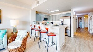 A modern kitchen with a central island, bar stools, and a dining area with a light-colored sofa. - Ocean-view 10th-floor condo with balcony, heated outdoor pool, hot tub, gym, W&D (Panama City Beach)