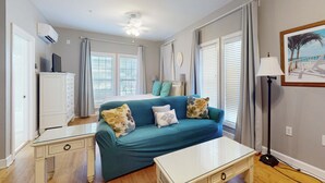 Ein Zimmer mit einem türkisfarbenen Sofa, einem weißen Tisch und einer Stehlampe. Es gibt ein Fenster mit Jalousien, einen Deckenventilator und ein gerahmtes Bild an der Wand.