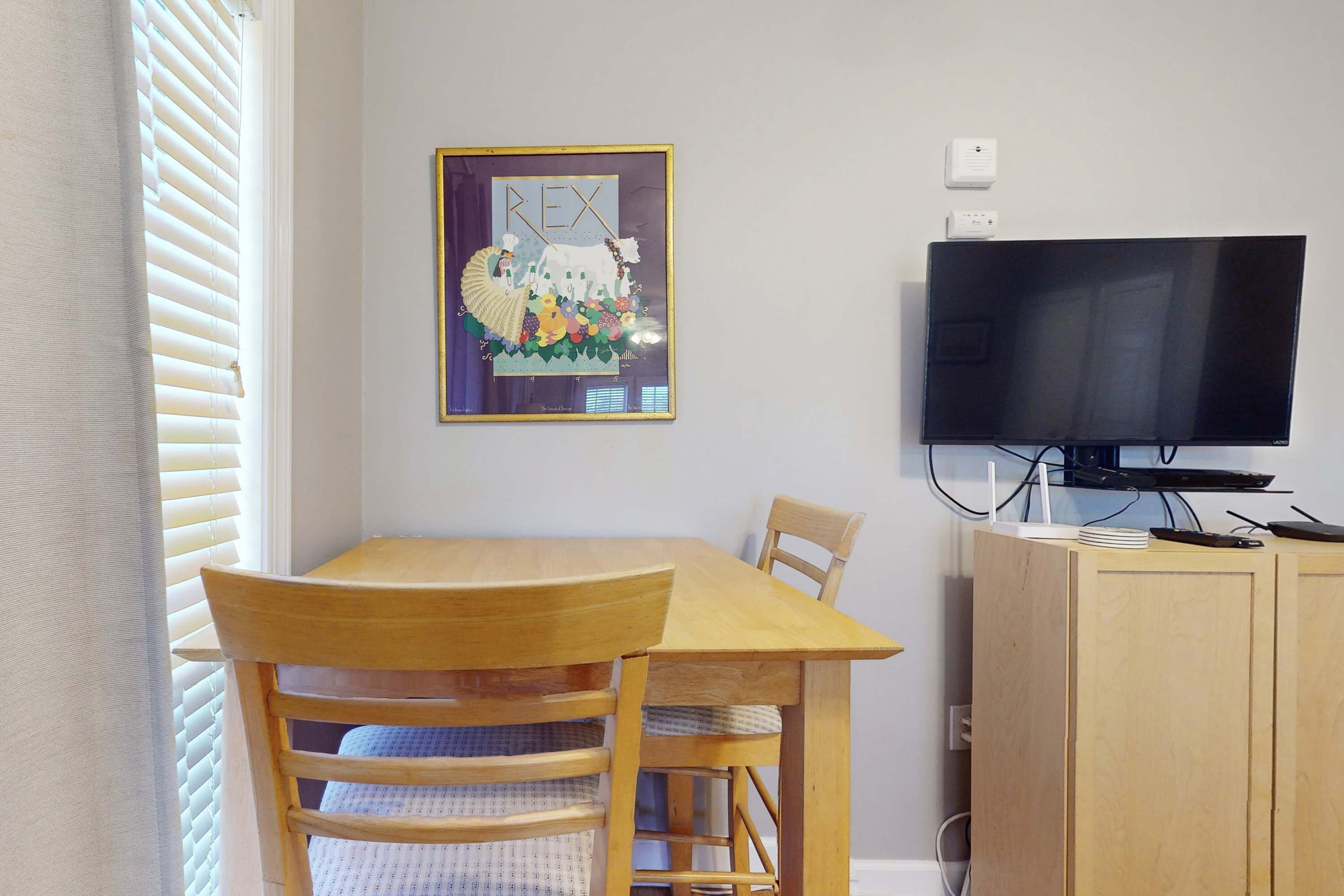 Ein Essbereich mit Holztisch und Stühlen, einem gerahmten Kunstwerk an der Wand, einem Fernseher auf einem Standfuß und einem Schrank mit Flachbildfernseher.