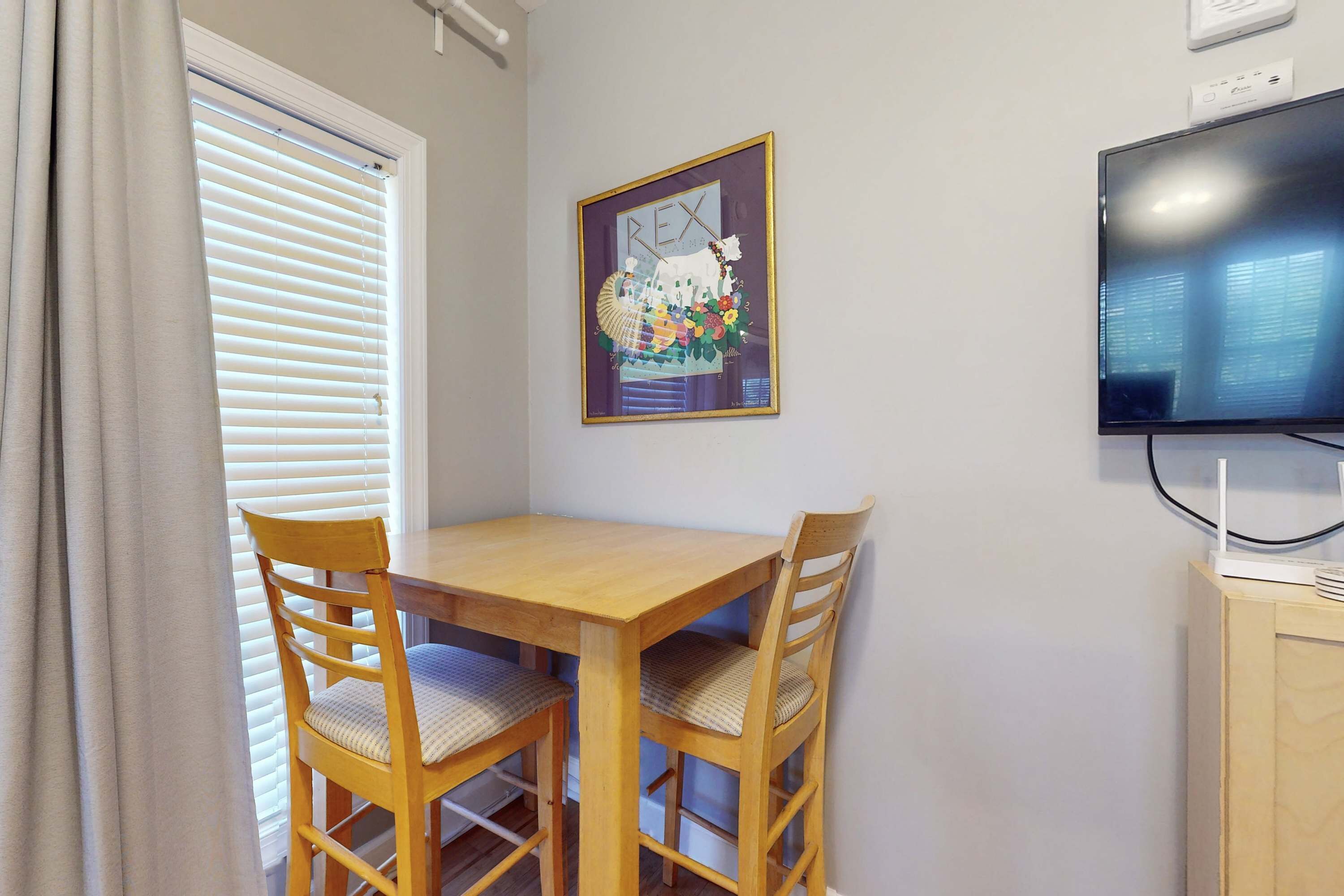 Un comedor con una mesa y sillas de madera, una obra de arte enmarcada en la pared, un televisor en la pared y una ventana con persianas.