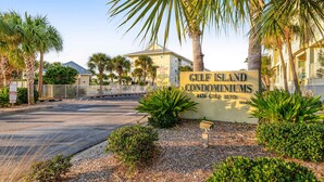En grind markerar ingången till Gulf Island Condominiums, med en tydlig väg som leder in, omgiven av palmer och välskött trädgård.