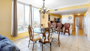 A dining area with a glass table and wicker chairs, a chandelier, and a view through large windows. - 3rd-Floor Gulf-Front Condo w/Balcony, Shared Hot Tub, Heated Pool, & Fast WiFi (Navarre Beach)