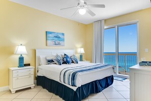 Ein Schlafzimmer mit einem Bett, zwei Nachttischen mit Lampen, einem Deckenventilator, einem Gemälde und einem Balkon mit Meerblick.