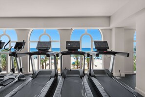 Ein Fitnessstudio mit Laufbändern und Trainingsgeräten, große Fenster mit Blick auf einen Strand und Palmen.