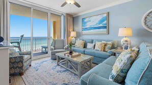 A room with a sofa, armchairs, a coffee table, a ceiling fan, a painting, a lamp, and a view of the ocean. - Enchanting oceanfront condo with pool, balcony, fitness area, & washer&dryer (Fort Walton Beach)