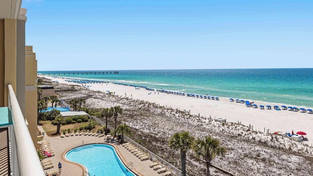 A beach view from a balcony with a pool, beach umbrellas, and a pier in the distance. - Enchanting oceanfront condo with pool, balcony, fitness area, & washer&dryer (Fort Walton Beach)