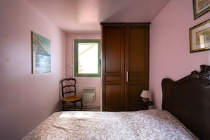 Un dormitorio con cama, una silla, un armario, una ventana y un cuadro colgado en la pared.