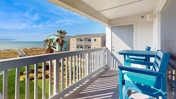 Un balcon avec vue sur la plage et l’océan, une balustrade blanche et des meubles bleus.
