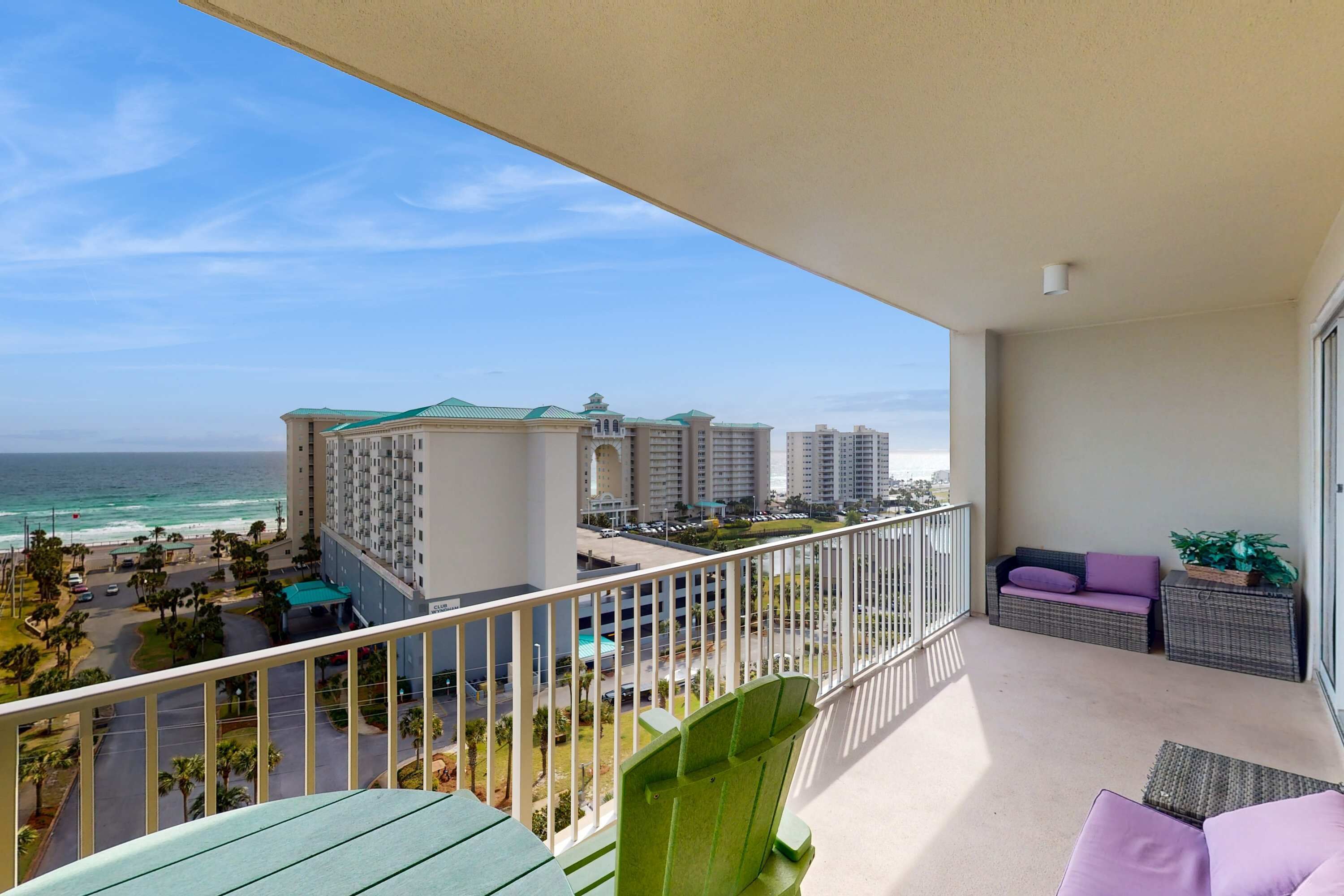 Uma varanda com vista para a praia e oceano, uma cadeira verde, um sofá roxo e uma mesa lateral de vime.
