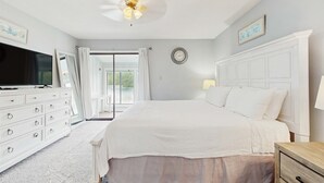 Un dormitorio con cama, cómoda, televisor, ventilador de techo, ventana con cortinas y vistas al exterior.