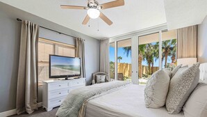Een slaapkamer met een bed, een televisie, een plafondventilator en uitzicht op palmbomen door het raam.