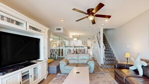 A spacious living room with a large flat-screen TV, a ceiling fan, a light-colored sofa, a coffee table, and a staircase leading to an upper level.