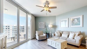 Amplia sala de estar con un ventanal grande, ventilador de techo, sofá color beige, mesa de centro blanca y una obra enmarcada en la pared.