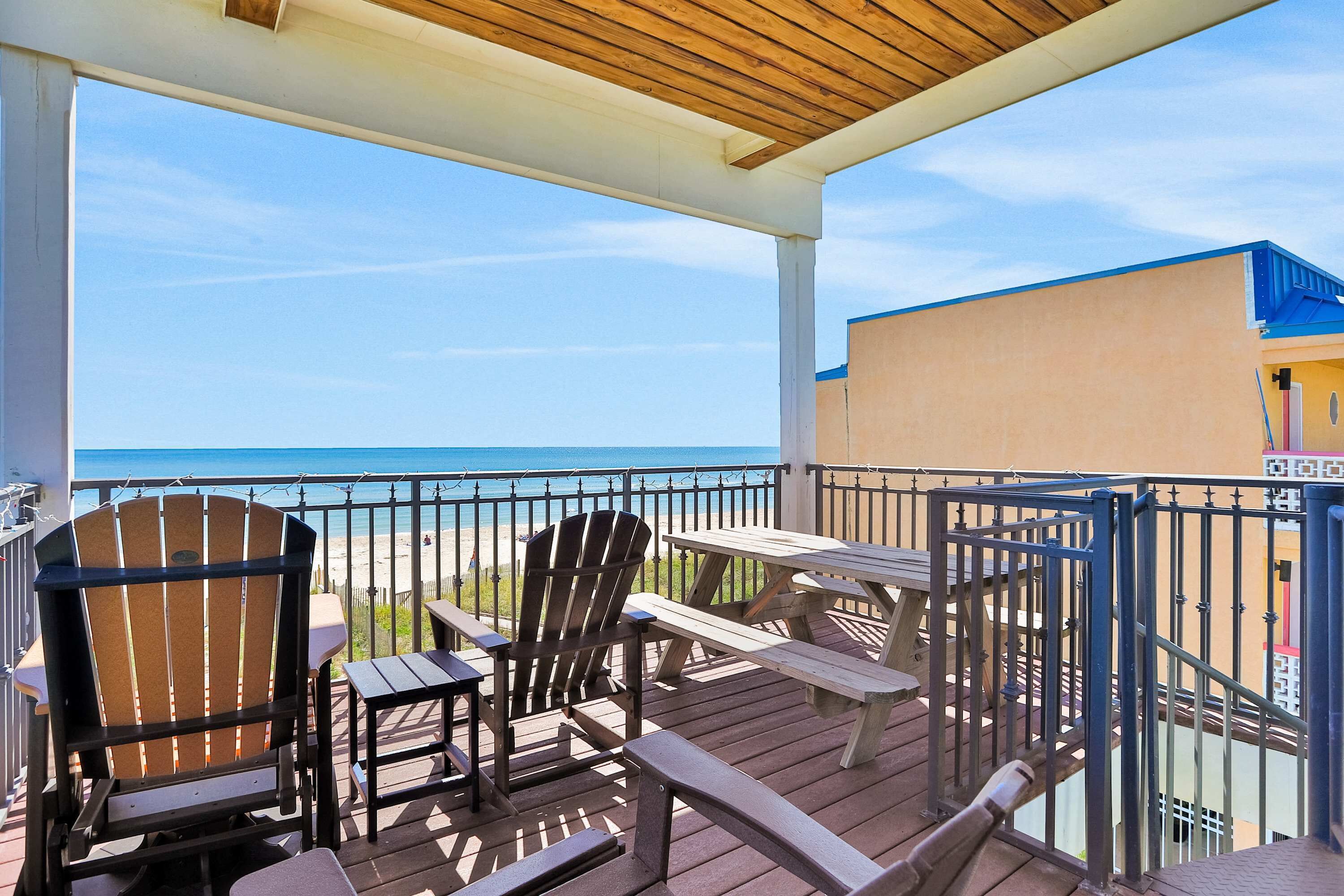 A wooden deck with Adirondack chairs and picnic tables overlooking a beach.