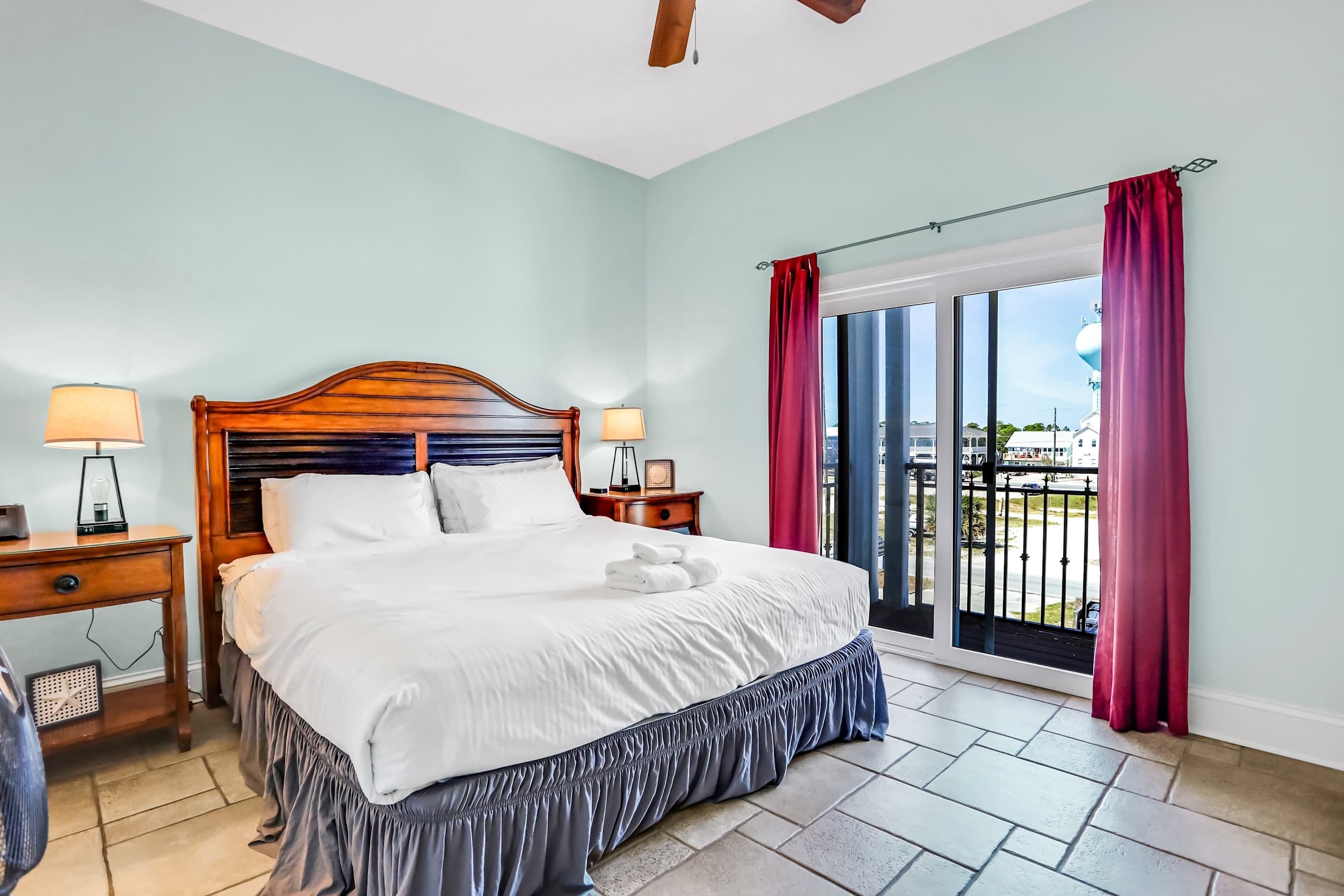 Habitación de hotel con una cama grande, dos mesitas de noche con lámparas y vistas a la ciudad desde un balcón con cortinas rojas.