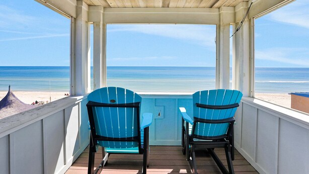 Varanda coberta com duas cadeiras Adirondack azuis e uma pequena mesa, com vista para a praia.