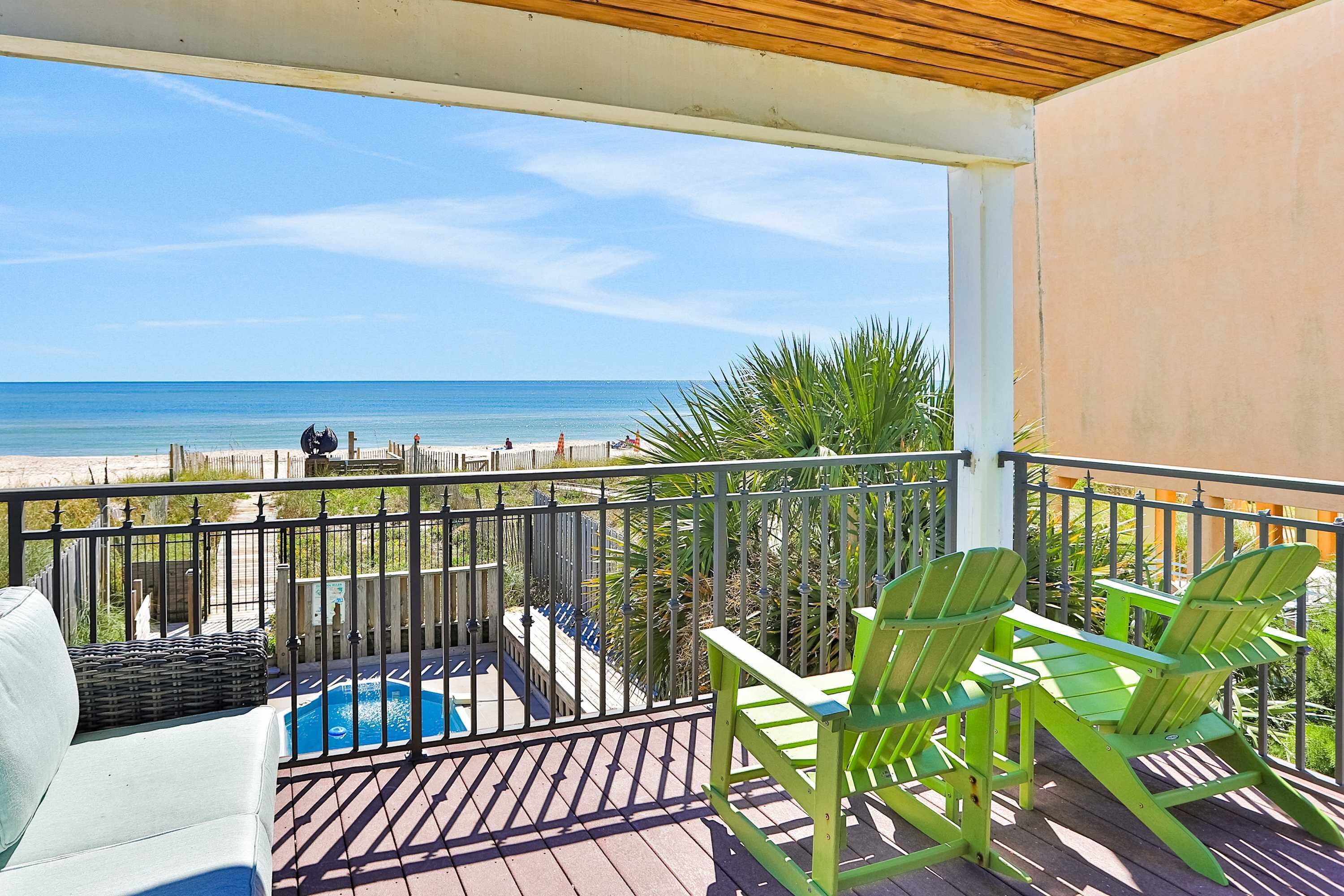 A balcony with a view of the beach, green Adirondack chairs, a pool, and a white couch.