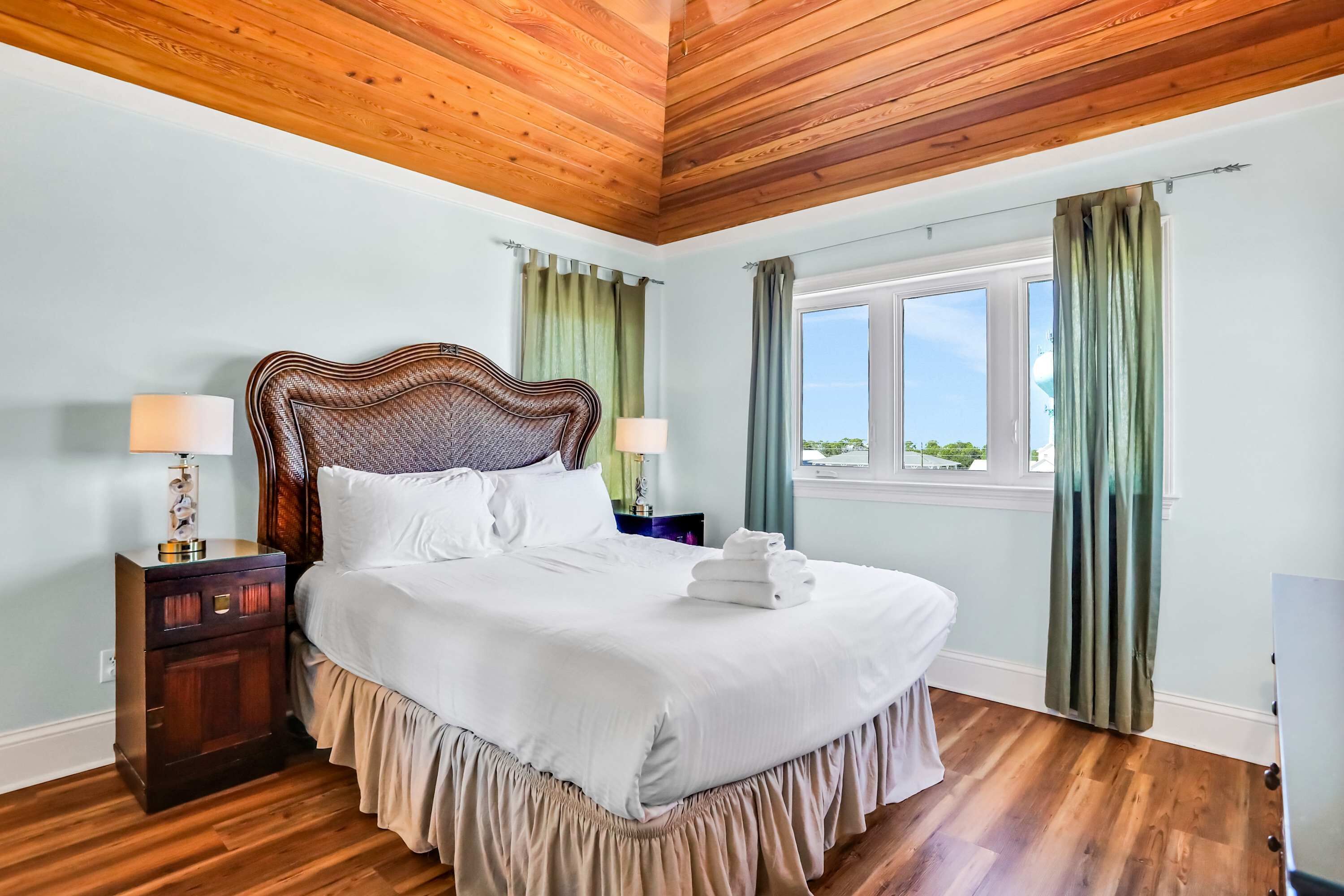 A neatly made bed with a wooden headboard, two bedside tables with lamps, a window with curtains, and a view of greenery outside.
