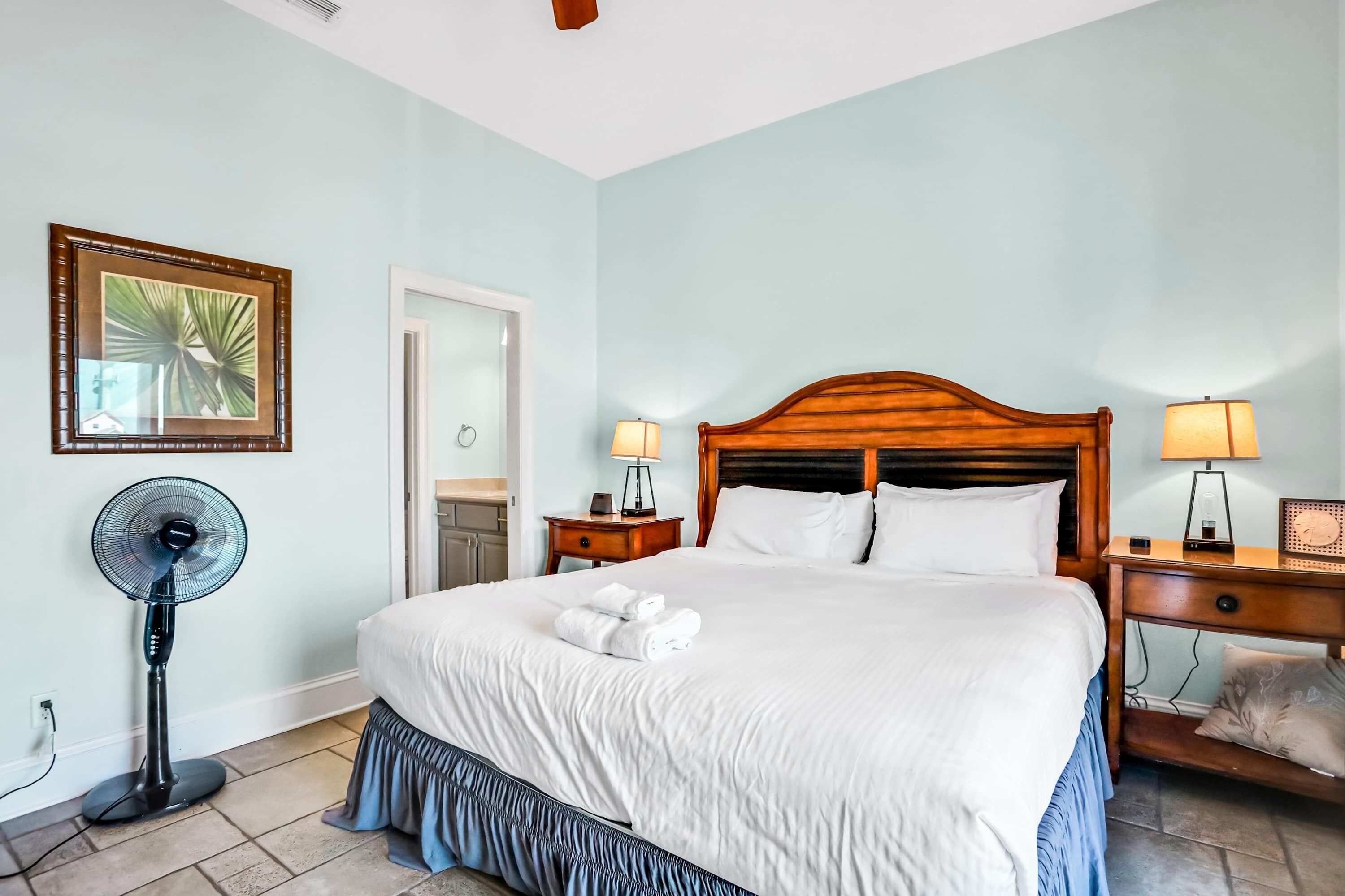 A neatly made bed with white linens and a blue skirted bed skirt, two bedside tables with lamps, a standing fan, and a framed picture on the wall.