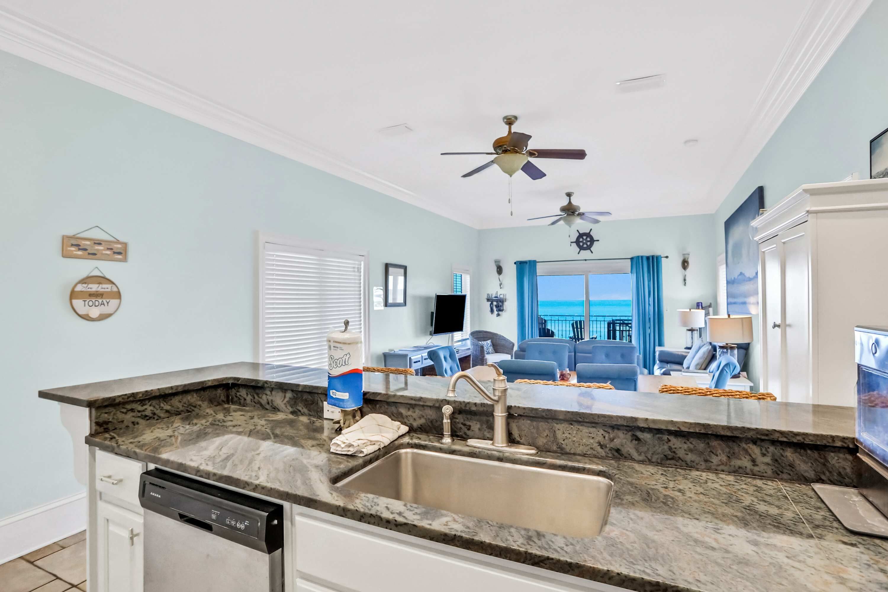 A kitchen with a granite countertop, stainless steel sink, and a dishwasher. There is a ceiling fan, a window with blue curtains, and a view of the ocean.