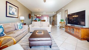 Ein geräumiges Wohnzimmer mit einem beigefarbenen Sofa, einem braunen Hocker, einem Deckenventilator, einem Fernseher und einer Küche im Hintergrund.