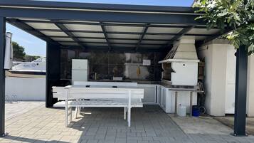 A covered outdoor kitchen area with a grill, a refrigerator, and a table.
