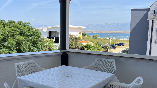 A balcony with a table and chairs, overlooking a scenic view of a beach and mountains. - Apartment Lavaran 4 in Vir (Vir)