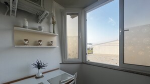 Een kamer met een witte tafel en stoelen, een klein plantje op de tafel, een raam met uitzicht op een gebouw, en een plank met decoratieve spullen.
