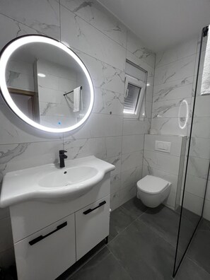 A modern bathroom with a white sink, a round mirror with a light, a toilet, and a shower area. - Apartment Lavaran 5 in Vir (Vir)