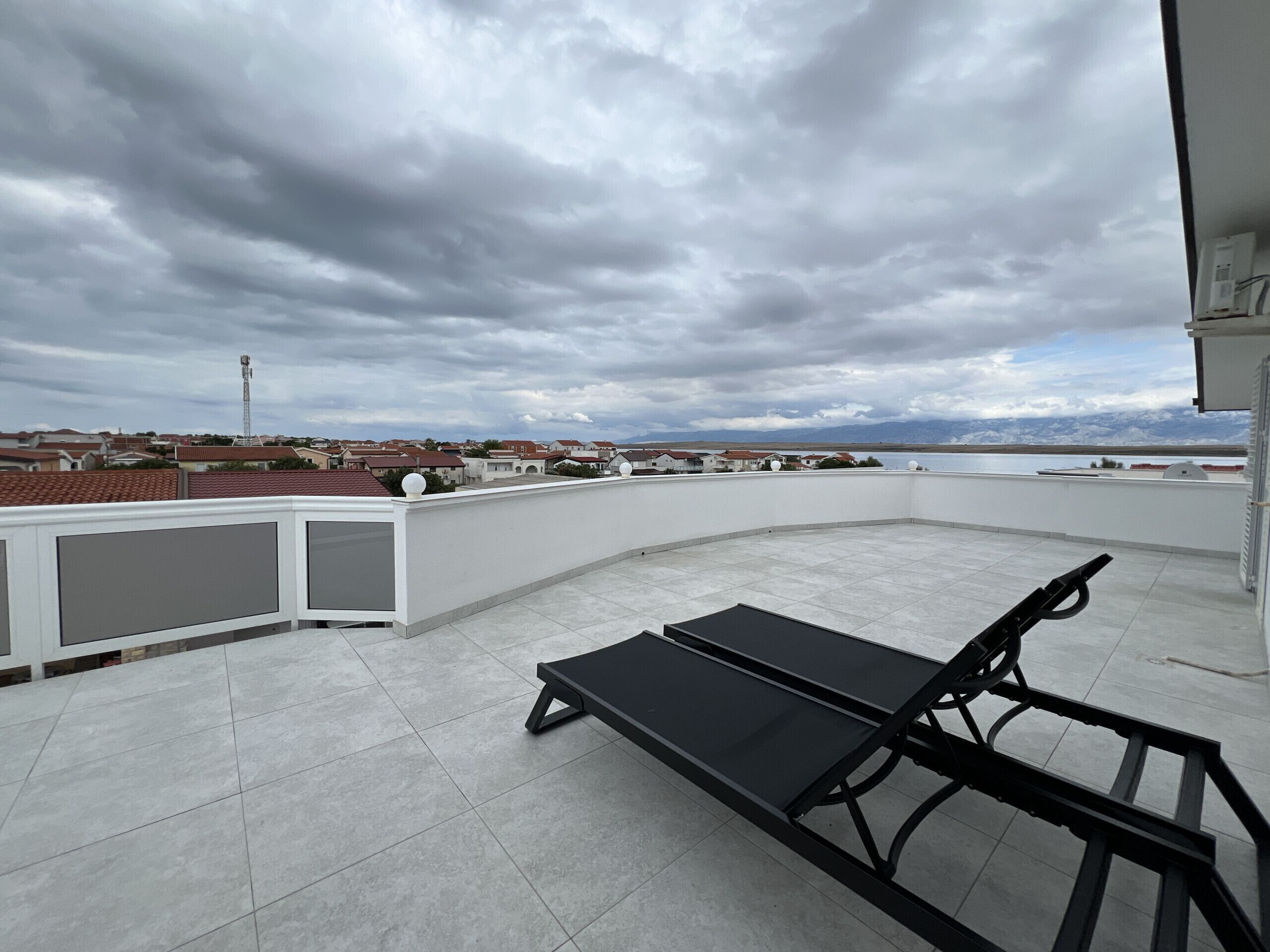 Una terraza en la azotea con una silla de lounge negra y una mesa, con vistas a un barrio residencial con casas y una cadena montañosa distante.