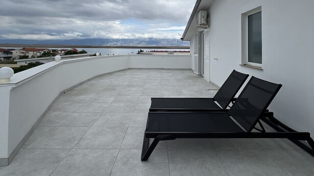 A rooftop terrace with a black lounge chair and a small table, overlooking a body of water and distant buildings. - Apartment Lavaran 6 in Vir (Vir)