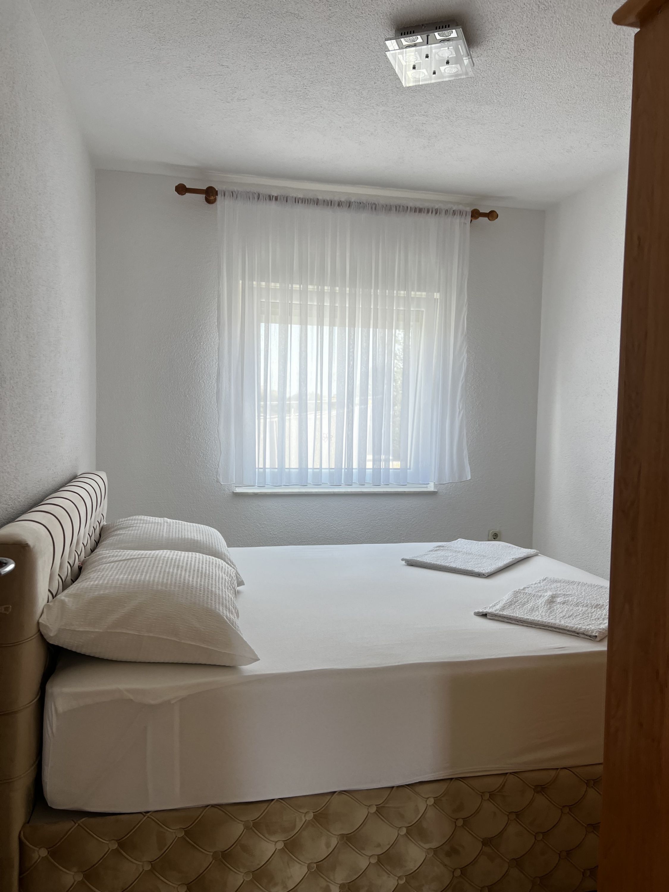 A bedroom with a bed, two pillows, a window with sheer curtains, and a ceiling light fixture.