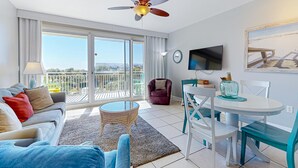 A modern living room with a sofa, a round coffee table, a dining table with chairs, a ceiling fan, a TV mounted on the wall, and a balcony with a view.