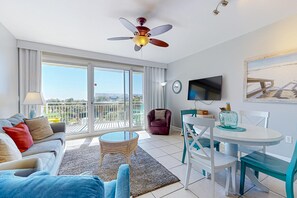 A modern living room with a sofa, a round coffee table, a dining table with chairs, a ceiling fan, a TV mounted on the wall, and a balcony with a view. - Elegant condo w sweeping gulf views, shared pools, snack bar & grill area (Destin)