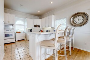 A kitchen with white cabinetry, a large island with bar stools, a clock on the wall, and a window with blinds. - Beachfront home with elevator, beach gear, on-site neiborhood pool, tennis & fitness room (St. George Island)