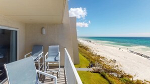Een balkon met twee stoelen en een tafel, met uitzicht op een strand met parasols en een pier.