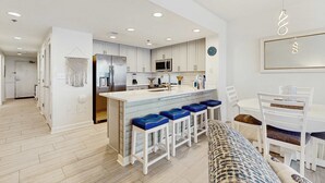 Een moderne keuken met witte kasten, een centraal eiland met blauwe barstoelen, en een eethoek met witte stoelen en een tafel.