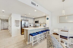 A modern kitchen with white cabinets, a central island with blue barstools, and a dining area with white chairs and a table. - Condo w beach access, gulf views & pool - snowbird friendly! (Destin)