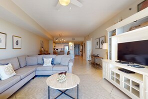 A spacious living room with a sectional sofa, a round coffee table, and a flat-screen TV on a white entertainment unit. - Magnificent waterfront condo w private balcony, shared pool, gym, & snack bar (Destin)