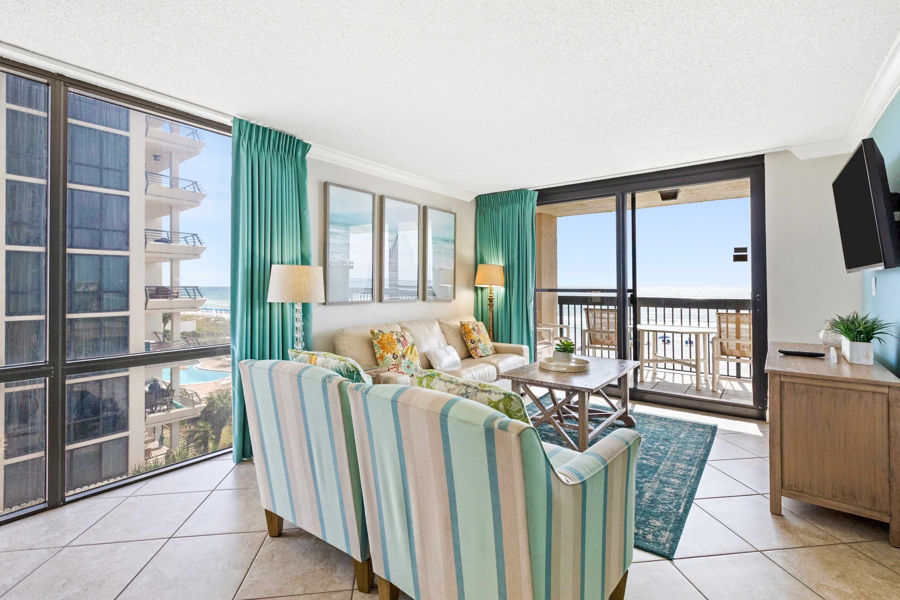 A spacious living room with a sofa, a coffee table, and a TV. Large windows offer a view of the pool and building.