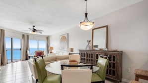 Een ruime woonkamer met een ronde glazen tafel, groene stoelen, een beige bank, een plafondventilator, een houten kast en een groot raam met uitzicht op de oceaan.