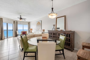 A spacious living room with a round glass table, green chairs, a beige sofa, a ceiling fan, a wooden cabinet, and a large window offering a view of the ocean. - Oceanfront Condo with Free WiFi, Central AC, W&D, Shared Hot Tub, Outdoor Pool, Gym (Pensacola Beach)