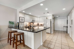 A modern kitchen with white cabinetry, a black countertop, and a central island with stools. - Oceanfront Condo with Free WiFi, Central AC, W&D, Shared Hot Tub, Outdoor Pool, Gym (Pensacola Beach)