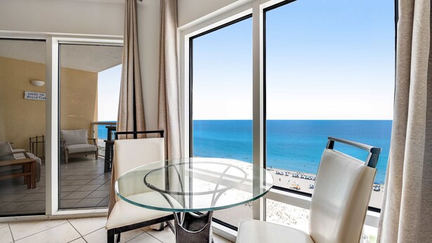 Een balkon met een glazen tafel en stoelen, met uitzicht op het strand en de oceaan.