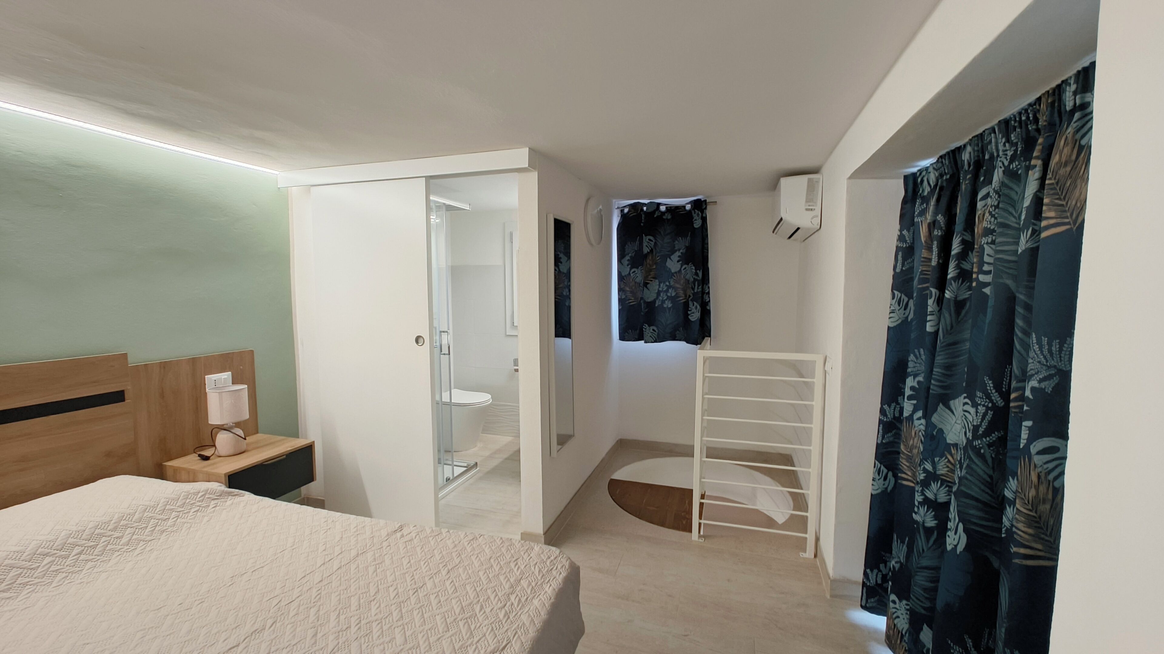 Ein Schlafzimmer mit Bett, Nachttisch mit Lampe, Kleiderschrank, Duschbereich und einem Fenster mit Vorhängen.