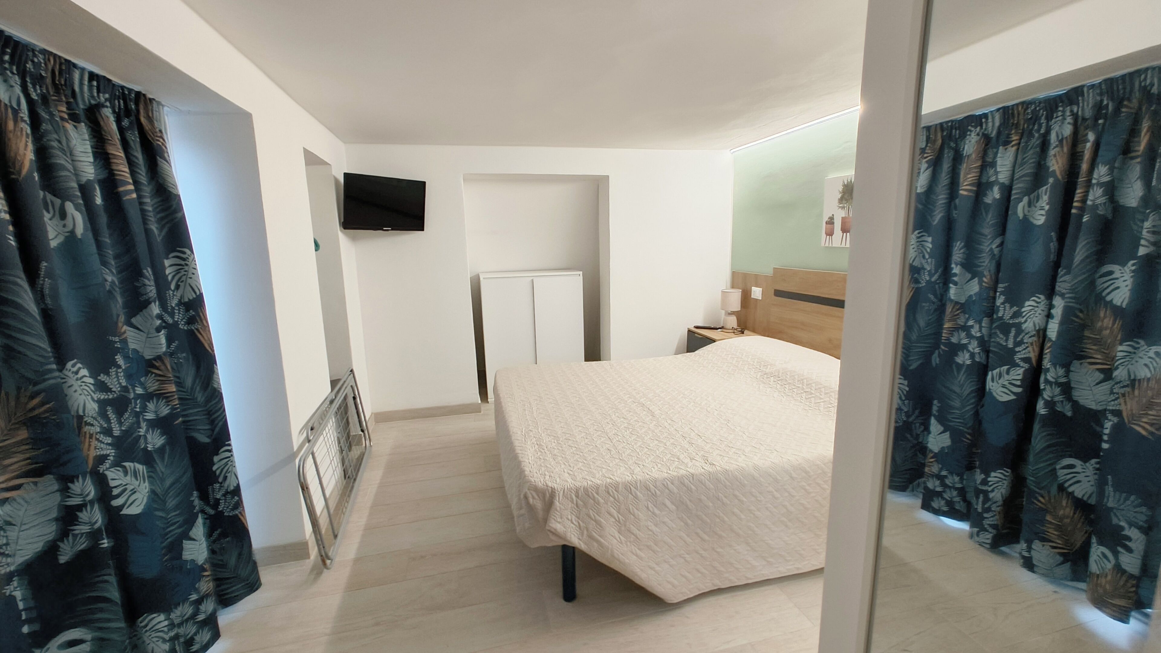 A hotel room with a bed, a TV mounted on the wall, and curtains with a tropical leaf pattern.