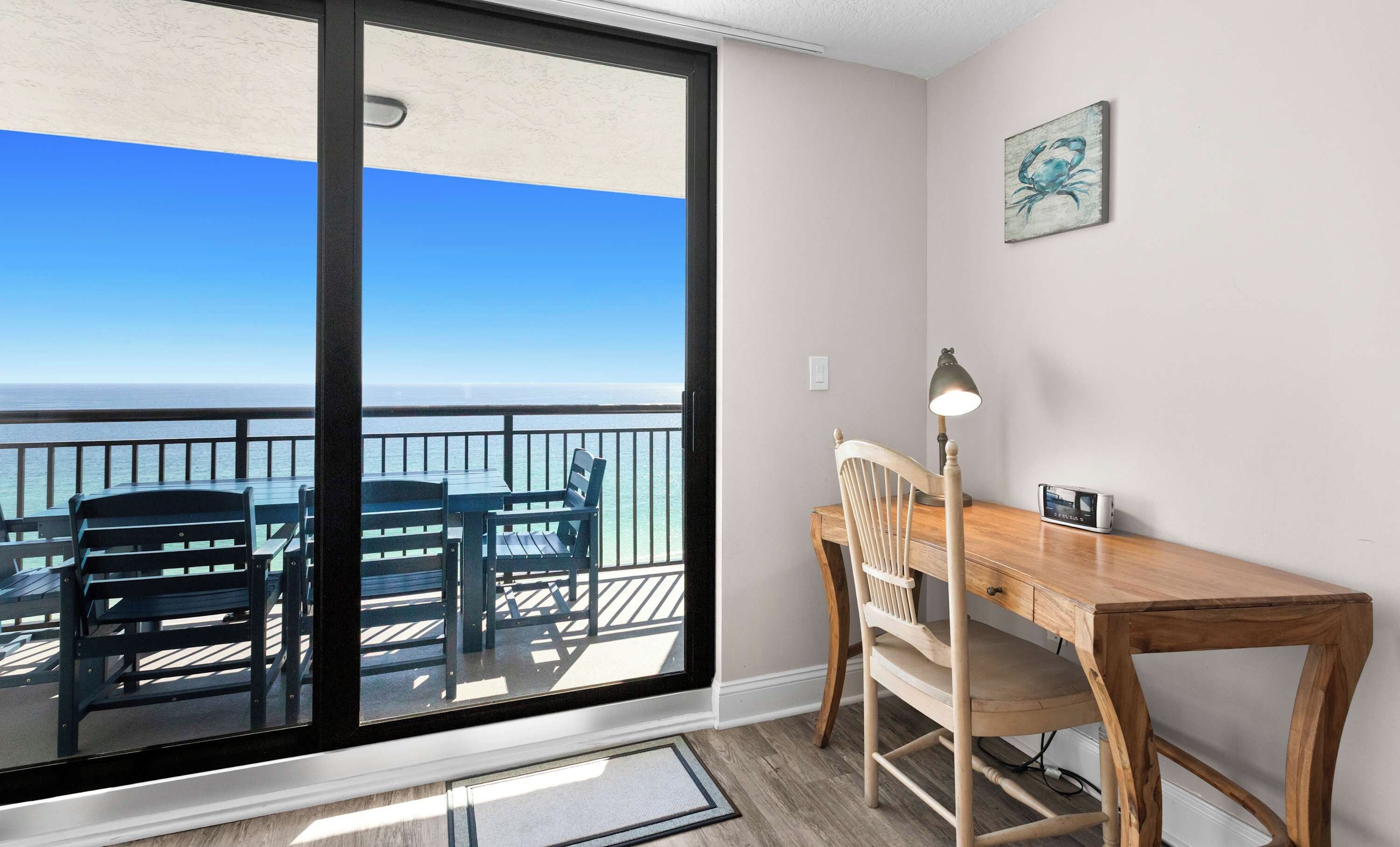 Een kamer met een grote schuifdeur die uitkomt op een balkon met uitzicht op de oceaan. Er is een houten bureau met een stoel en een lamp.