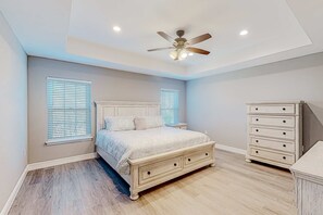 Ein Schlafzimmer mit Bett, Kommode, Deckenventilator und zwei Fenstern mit Jalousien.